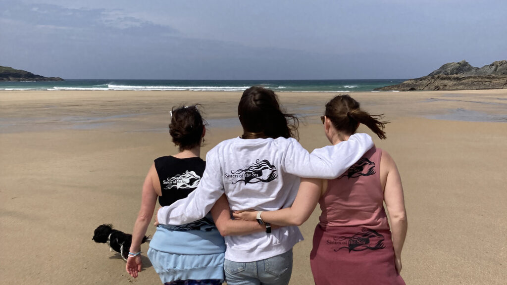 Three sisters of the sea from behind all linked with arms on the beach in front of the ocean.