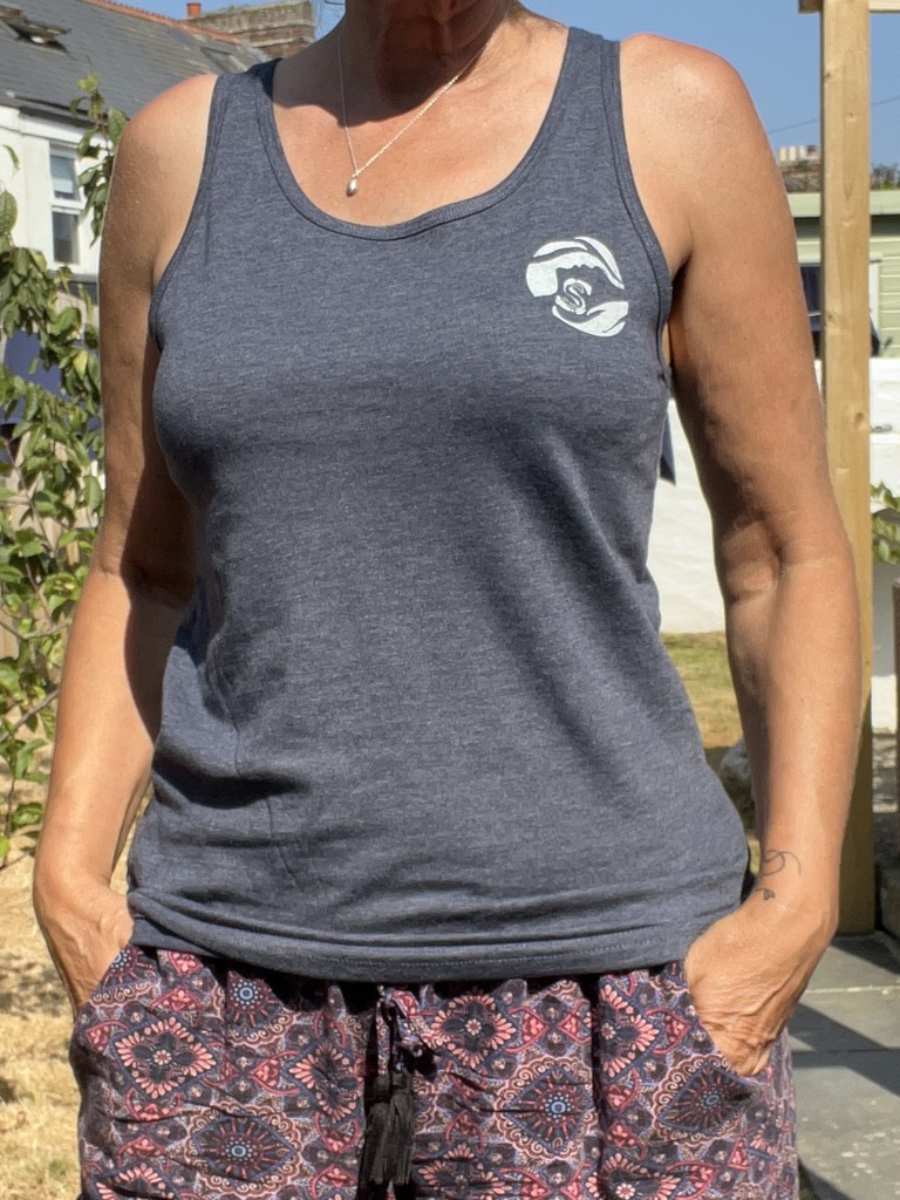 A close up of a woman standing in a garden wearing a blue Sisters of the Sea vest with a round logo on the left chest in white.