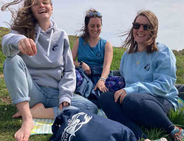 three women sitting on the grass with a navy blue sisters of the sea bag in front of the them, they are laughing.