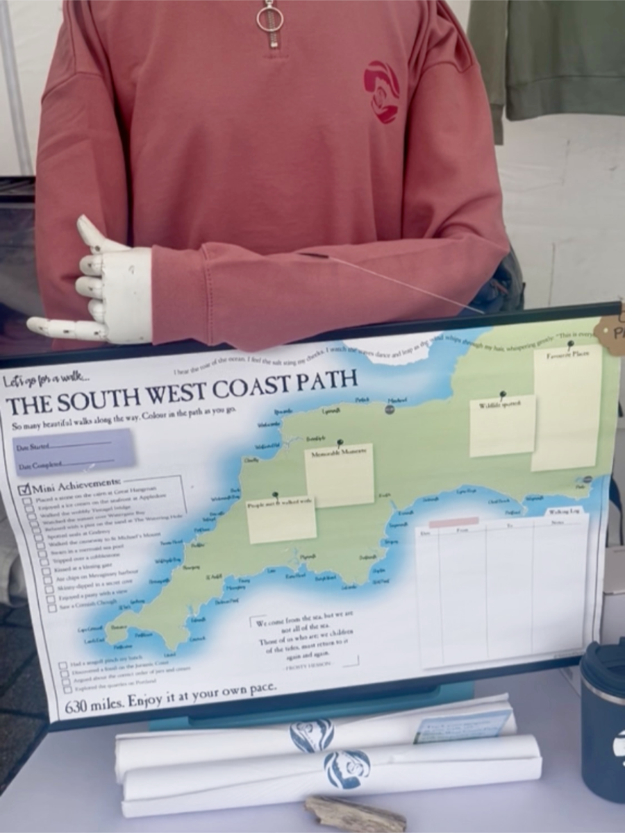 A photo of a manikin wearing a pink Sisters of the Sea afterglow quarter zip sweatshirt. It is holding a large poster with a map of Devon and Cornwall on it, detailing the South West Coast Path all the way around. There are rolled posters on the table in front of it.