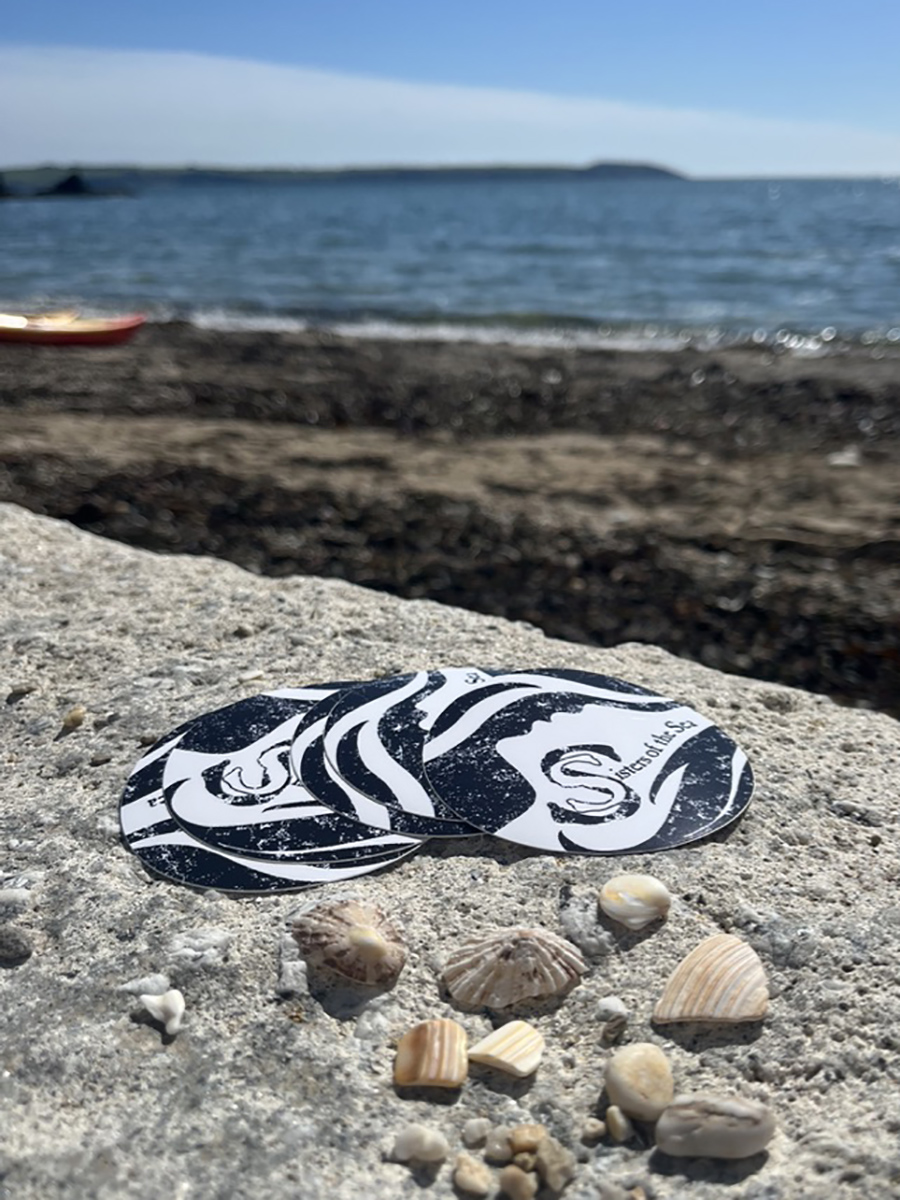 A pile of Sisters of the Sea stickers in black and white, in front of twinkling blue sea.