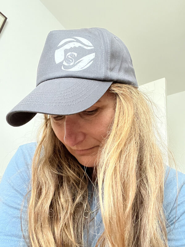 Model with long hair looking down, wearing a grey cap with white Sisters of the Sea logo.