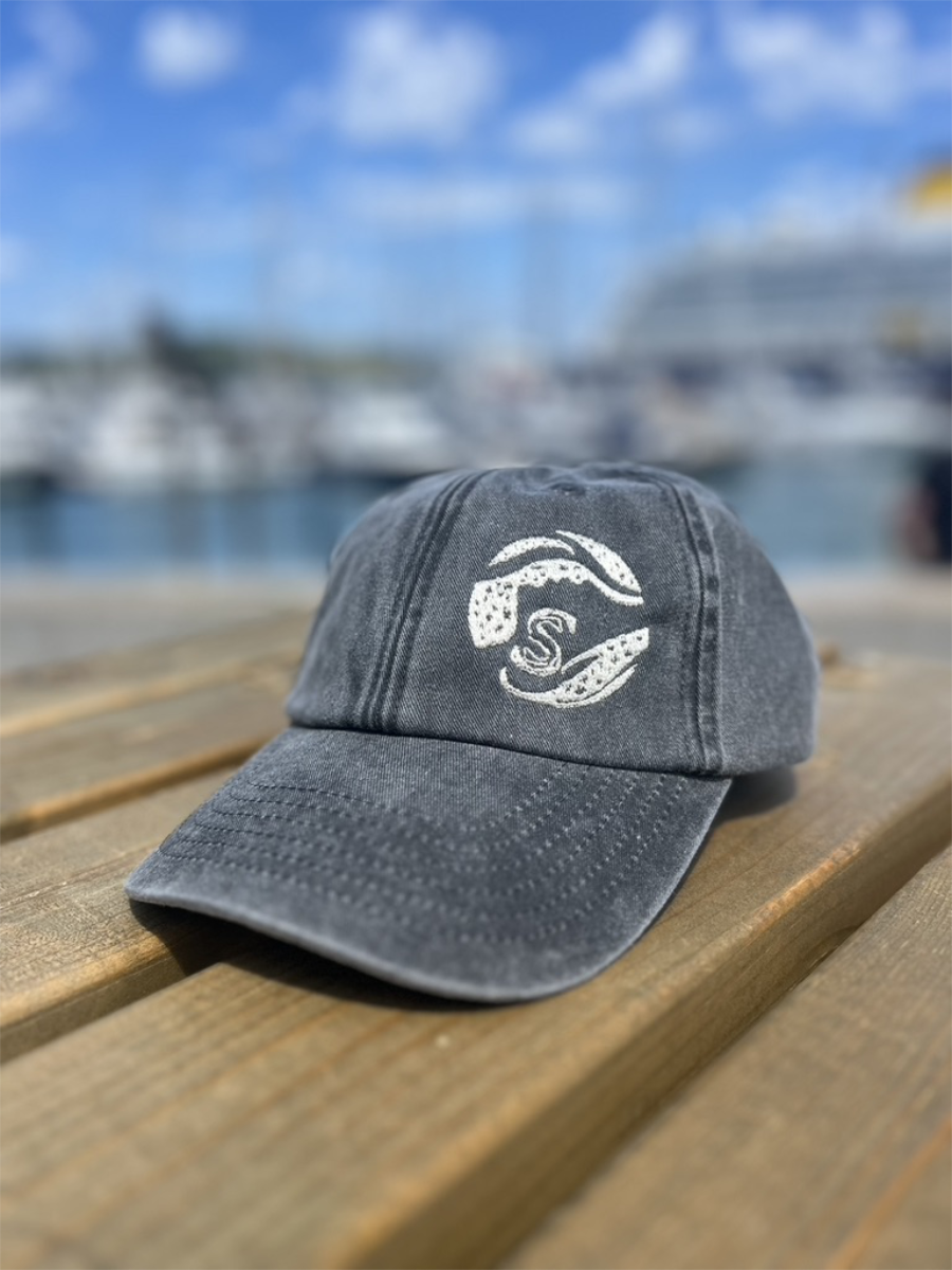 A black/grey Sisters of the Sea cap sitting on top of a wooden table with a harbour in the background.