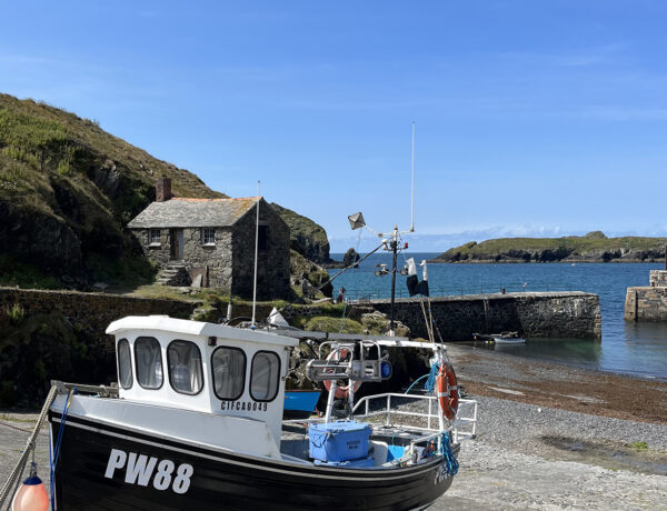 A fishing boat in the foreground with Mullion harbour behind. The harbour walls sit in front of beautiful blue seas and skies.