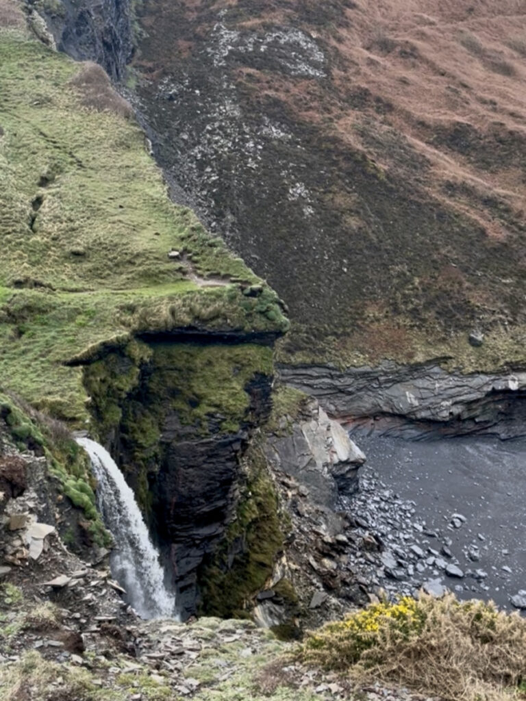 A photo of Pentargon waterfall near Boscastle in front of the heather covered cliffs and the beach below.