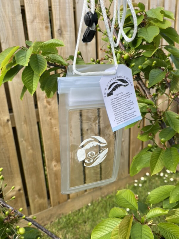 A waterproof phone pouch, which is transparent with white edges and a white Sisters of the Sea logo. There is a tag on the pouch with advice for use, and behind is a wood fence, with green leaves around.