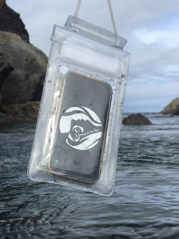 A waterproof phone pouch, which is transparent with white edges and a white Sisters of the Sea logo. It is hanging over the sea with a blue sky behind and rocks either side.