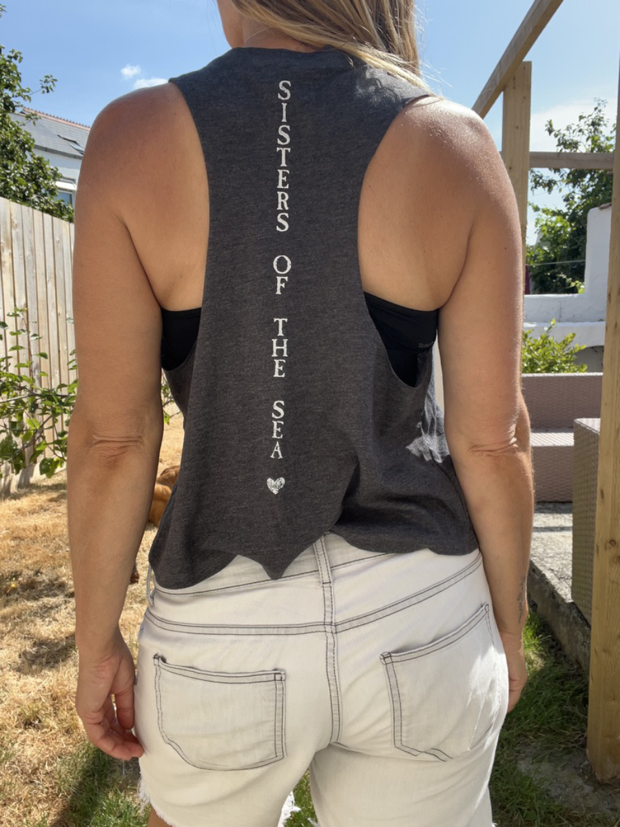 A close up photo of a woman from behind wearing a dark grey breezy tank vest with Sisters of the Sea written in white vertically down the back. She has her arms up tying her hair and there is a garden in the background.