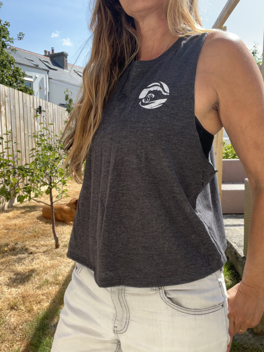 A close up photo of a woman wearing a dark grey breezy tank vest with a white circle Sisters of the Sea logo on the chest. There is a garden in the background and a dog lazing in the sun.
