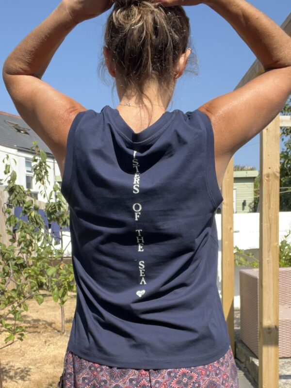 A close up photo of a woman from behind wearing a navy blue boxy vest with Sisters of the Sea written in white vertically down the back. She has her arms up tying her hair and there is a garden in the background.