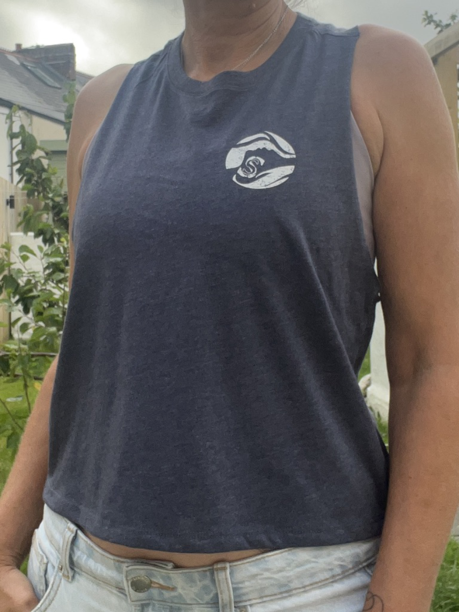 A close up photo of a woman wearing a navy breezy tank vest with a white circle Sisters of the Sea logo on the chest. There is a garden and tree in the background.