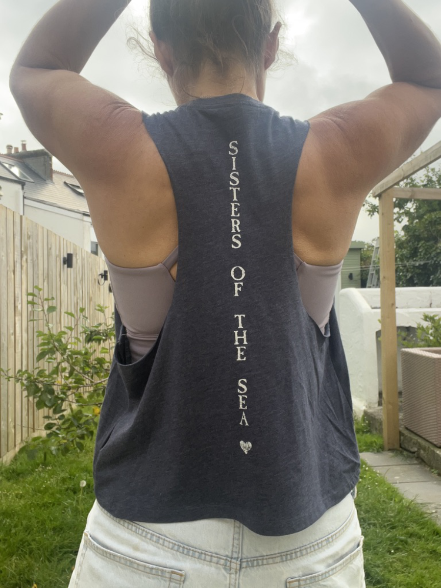 A close up photo of a woman from behind wearing a navy breezy vest with Sisters of the Sea written in white vertically down the back. She has her arms up tying her hair and there is a garden in the background.