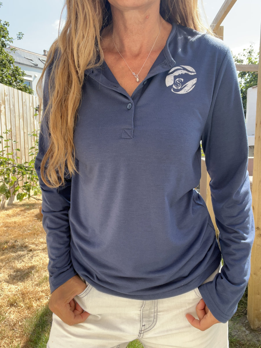 A photo of a woman wearing a navy blue Sisters of the Sea long sleeve henley top with a white round logo on the left chest and buttons down the front. There is a garden in the background.
