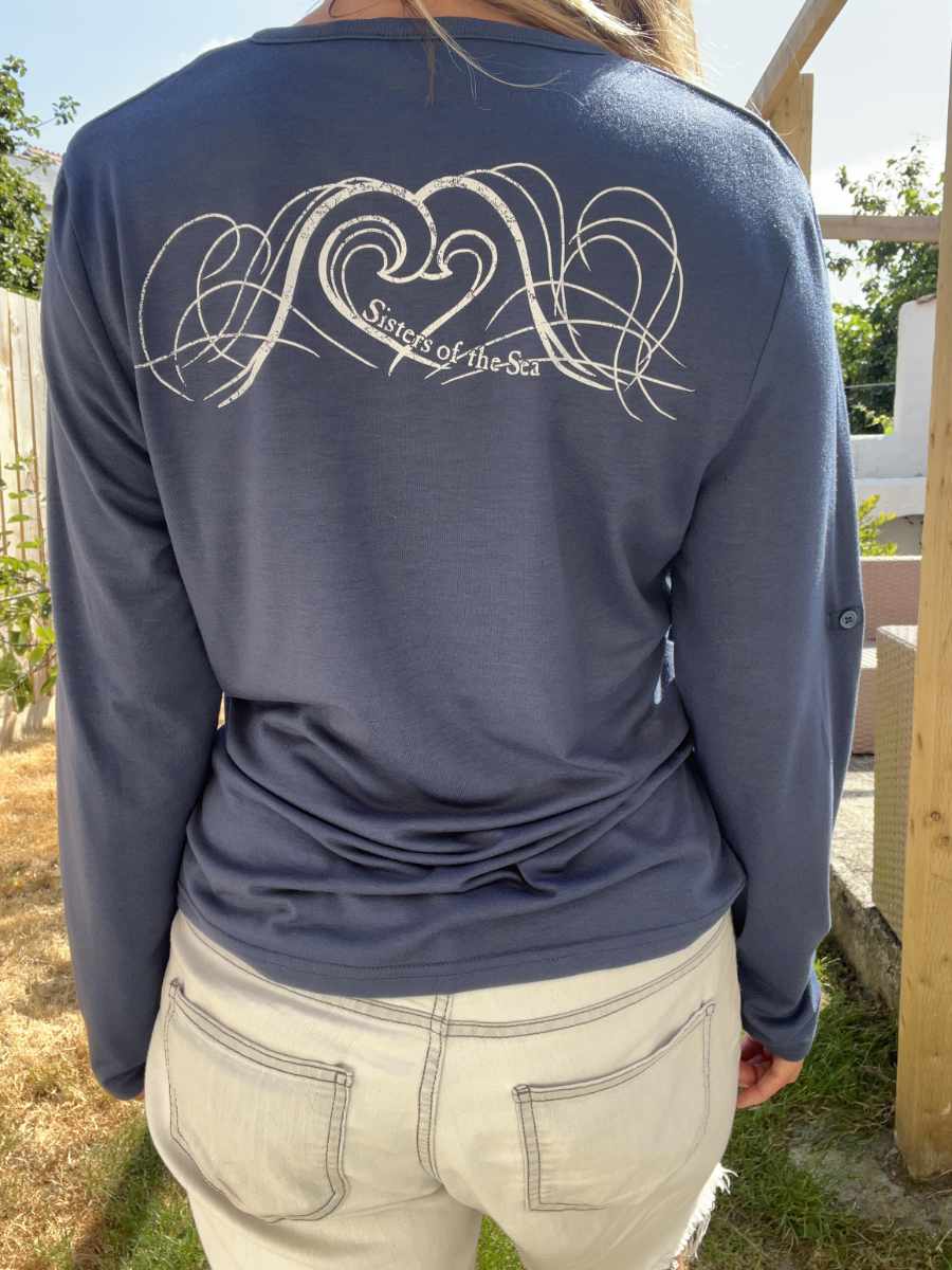 A photo of a woman from behind wearing a navy blue Sisters of the Sea long sleeve henley top with a logo showing waves that crash together to make a heart shape. There is a garden in the background.