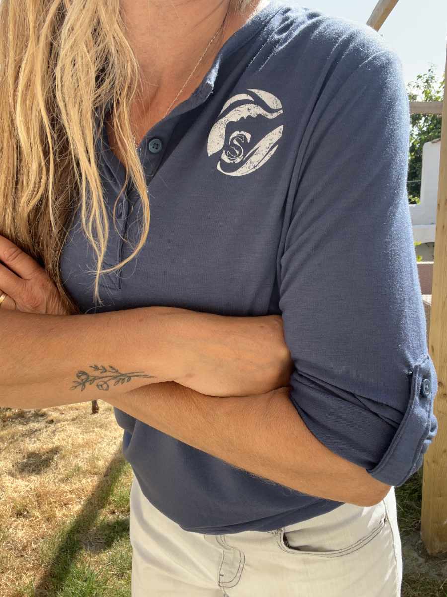 A photo of a woman with her arms crossed wearing a navy blue Sisters of the Sea long sleeve henley top with a white logo on the chest. She has long brown hair falling to one side and there is a garden in the background.