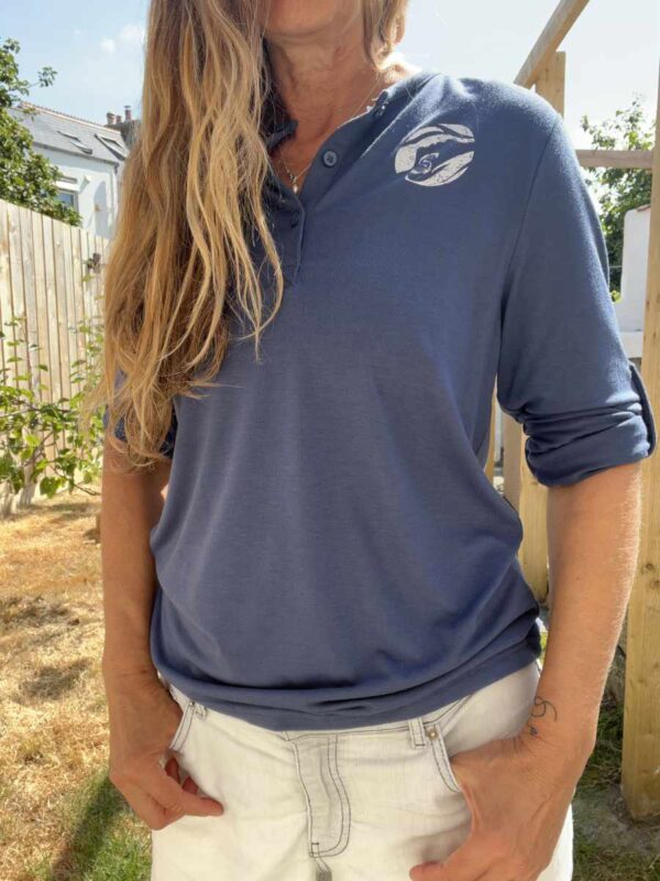 A photo of a woman wearing a navy blue Sisters of the Sea long sleeve henley top with a white round logo on the left chest and buttons down the front. There is a garden in the background.