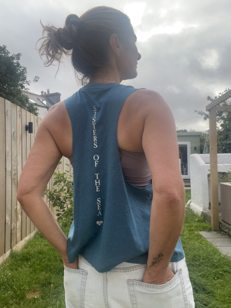 A close up photo of a woman from behind wearing an ocean teal breezy vest with Sisters of the Sea written in white vertically down the back. She has her arms up tying her hair and there is a garden in the background.
