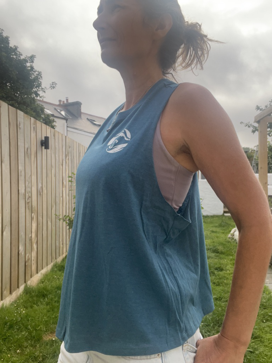 A close up photo of a woman wearing an ocean teal breezy tank vest with a white circle Sisters of the Sea logo on the chest. There is a garden in the background.