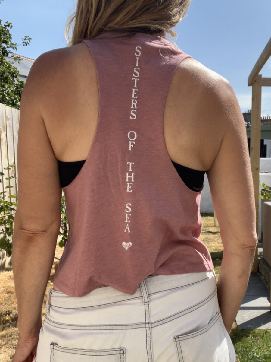 A close up photo of a woman from behind wearing a pink breezy vest with Sisters of the Sea written in white vertically down the back. She has her arms up tying her hair and there is a garden in the background.