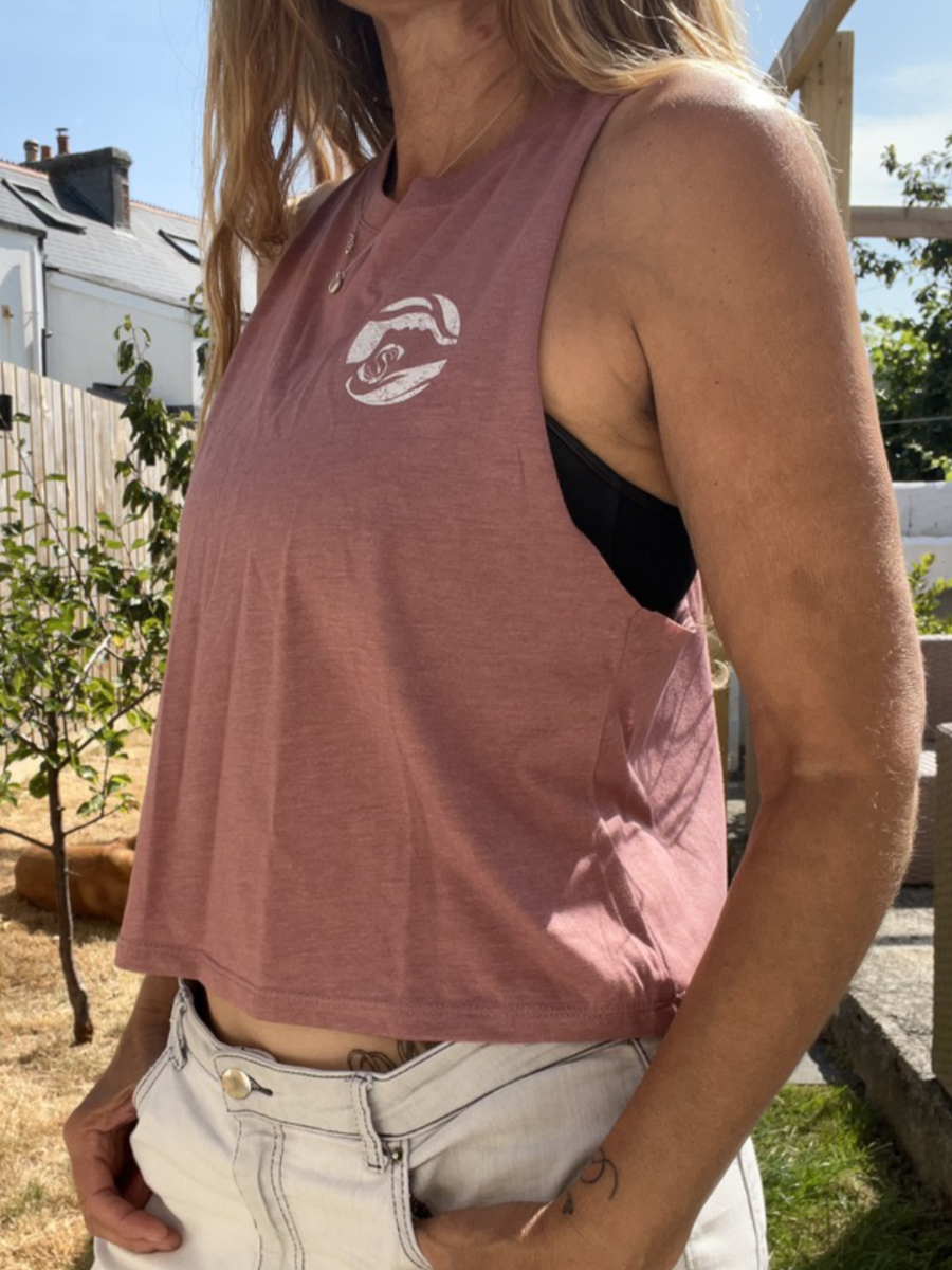 A close up photo of a woman wearing a pink breezy tank vest with a white circle Sisters of the Sea logo on the chest. There is a garden in the background and a dog lazing in the sun.