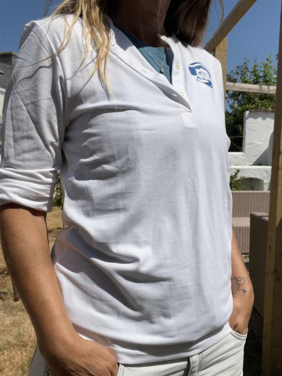 A photo of a woman wearing a white Sisters of the Sea long sleeve henley top with a blue round logo on the left chest. There are buttons down the front that are open and she has a teal vest underneath. Her sleeves are rolled up and there is a garden in the background.