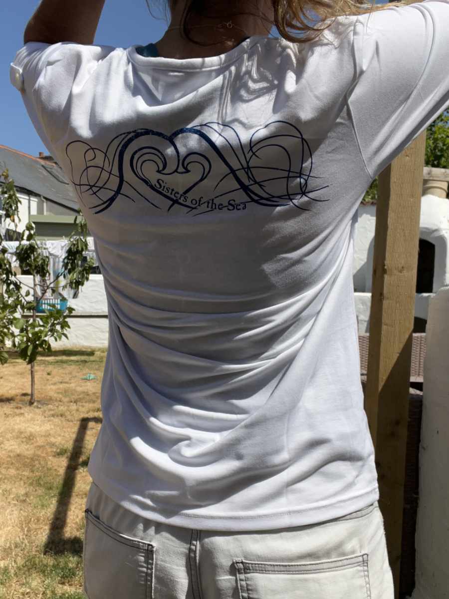 A photo of a woman from behind wearing a white Sisters of the Sea long sleeve henley top with a blue logo showing waves that crash together to make a heart shape. There is a garden in the background with a small tree and a pergola.