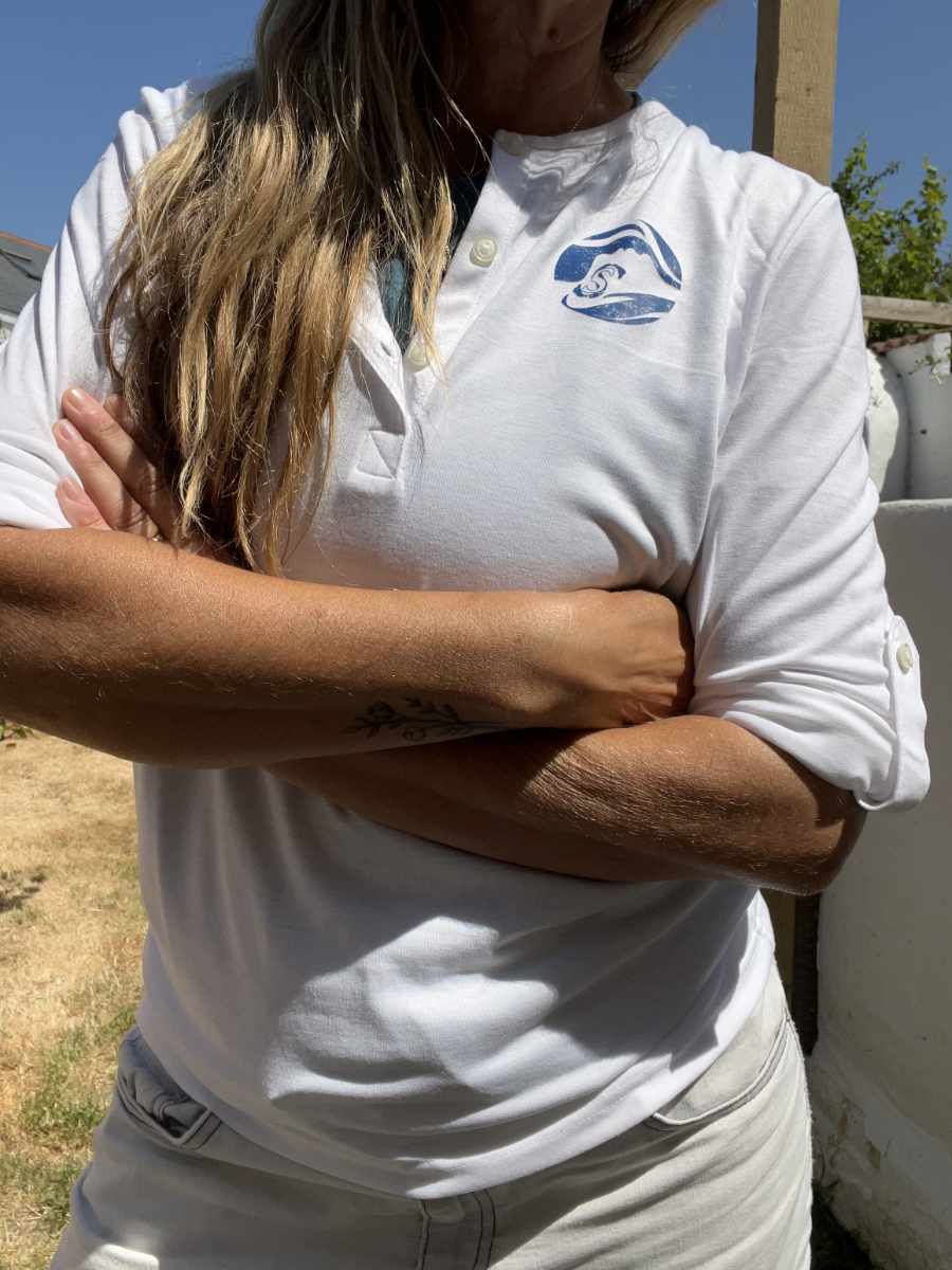 A photo of a woman wearing a white Sisters of the Sea long sleeve henley top with a blue round logo on the left chest. There are buttons down the front that are open and she has a teal vest underneath. Her sleeves are rolled up and she has her arms folded.