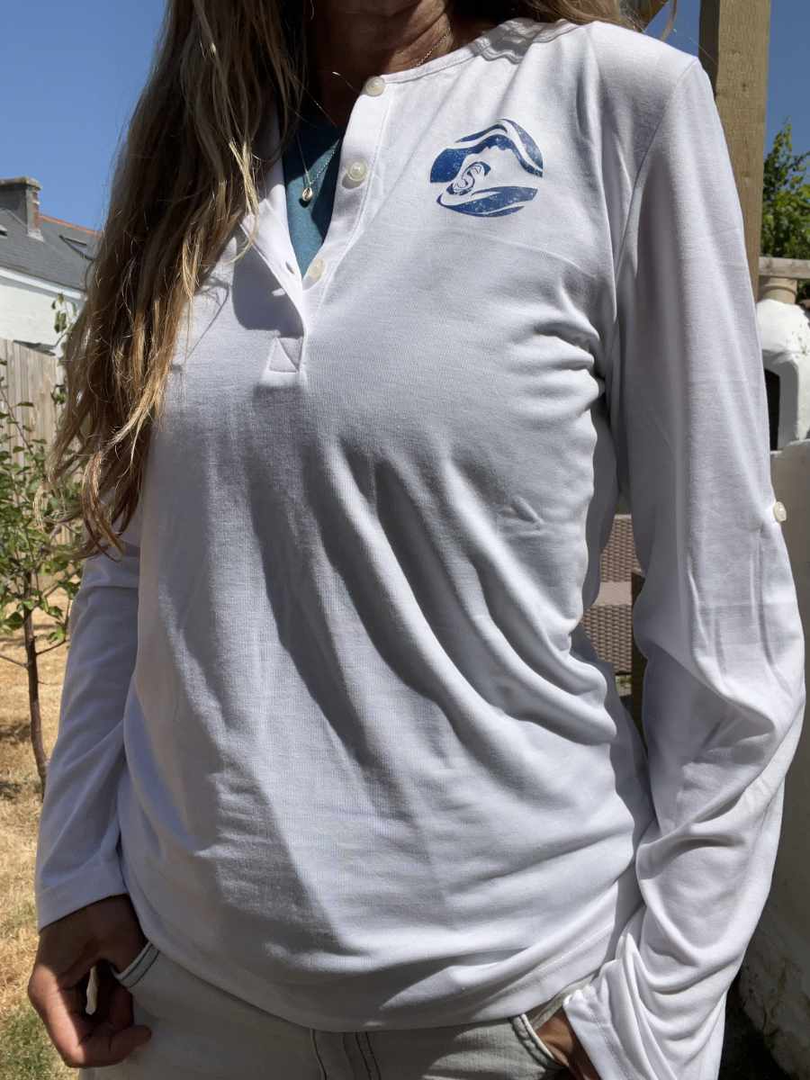 A photo of a woman wearing a white Sisters of the Sea long sleeve henley top with a blue round logo on the left chest. There are buttons down the front that are open and she has a teal vest underneath. Her hair falls onto her right shoulder and there is a garden in the background.