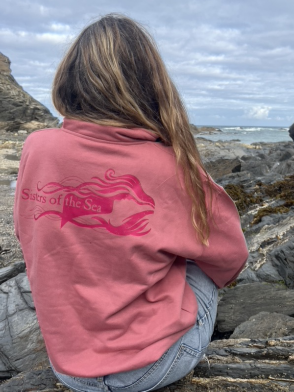 A photo of a woman sitting on a rock looking out to sea. She is wearing a pink sweatshirt with a bright pink Sisters of the Sea logo across the back. Her brown hair falls over her right shoulder and she wears blue denim shorts.