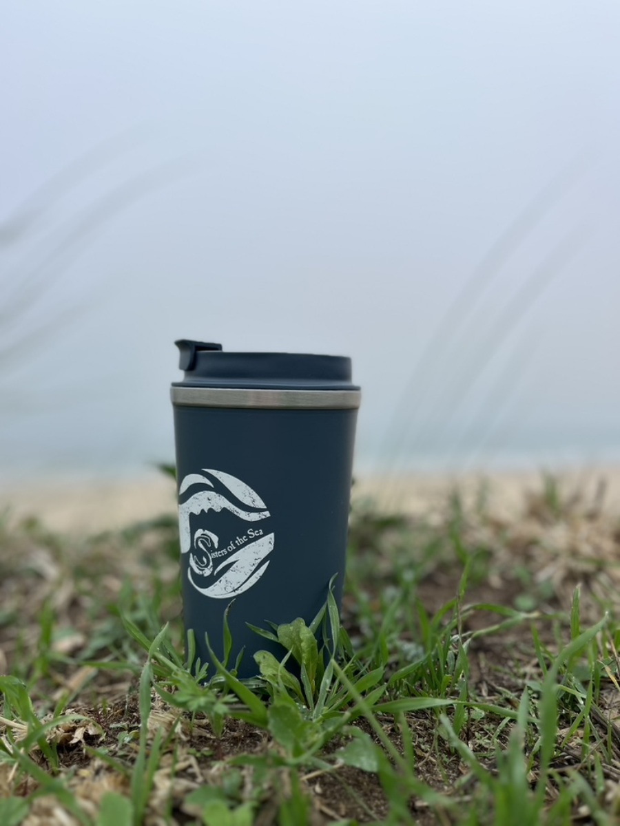 A dark blue travel mug with a white Sisters of the Sea logo. It sits on a grassy headland with a sandy beach behind it and the sea in the distance.