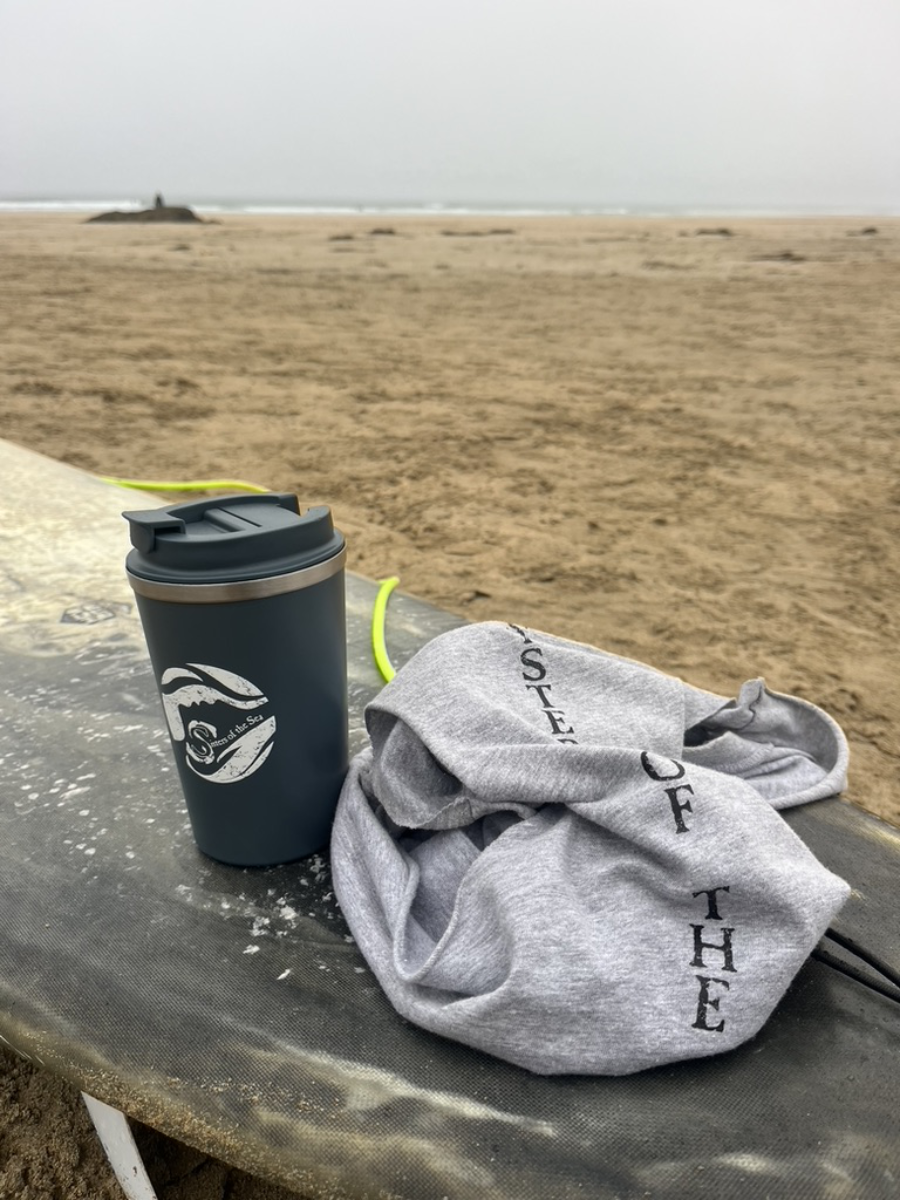 A photo of a dark blue travel mug sitting on top of a grey surfboard, with a grey vest beside it. Behind is sand and in the distance you can see the sea.