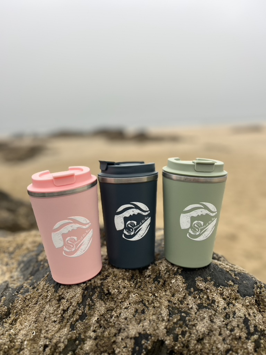 A photo of three travel mugs sitting on a rock on a sandy beach with the sea in the background. They are blue, green and pink and all have a white Sisters of the Sea logo on them.