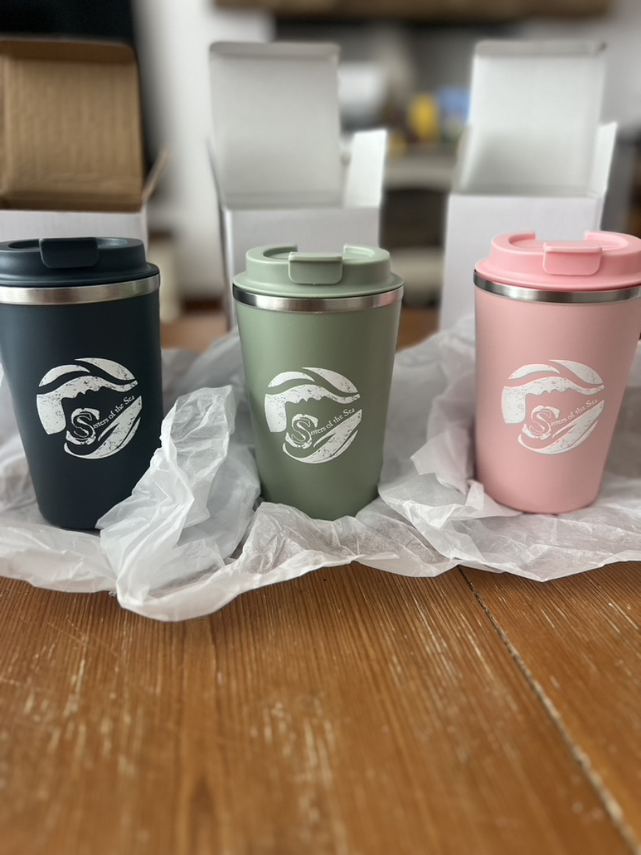 A photo of three travel mugs sitting on tissue paper with boxes behind. They are blue, green and pink and all have a white Sisters of the Sea logo on them.