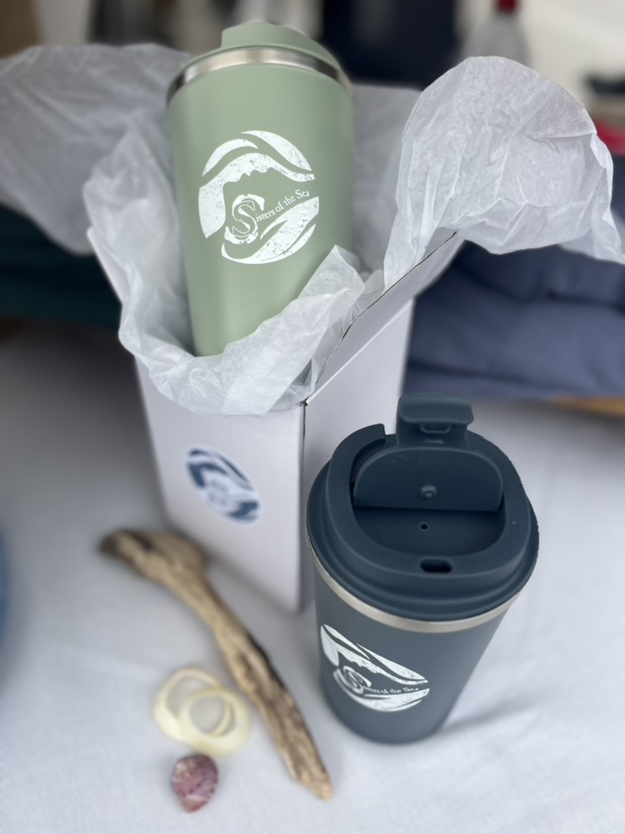 A green travel mug half inside a white box with tissue paper around it. Next to it on a white table is a blue travel mug and a few shells and driftwood. Both have a white Sisters of the Sea logo on them.