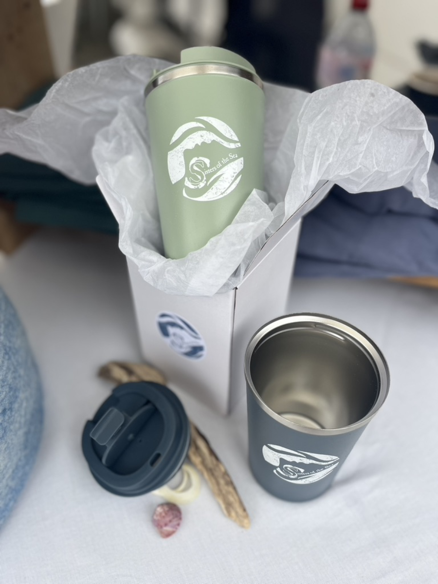A green travel mug half inside a white box with tissue paper around it. Next to it on a white table is a blue travel mug with the lid off. You can see the inside is stainless steel. Also on the table are a few shells and driftwood. Both have a white Sisters of the Sea logo on them.