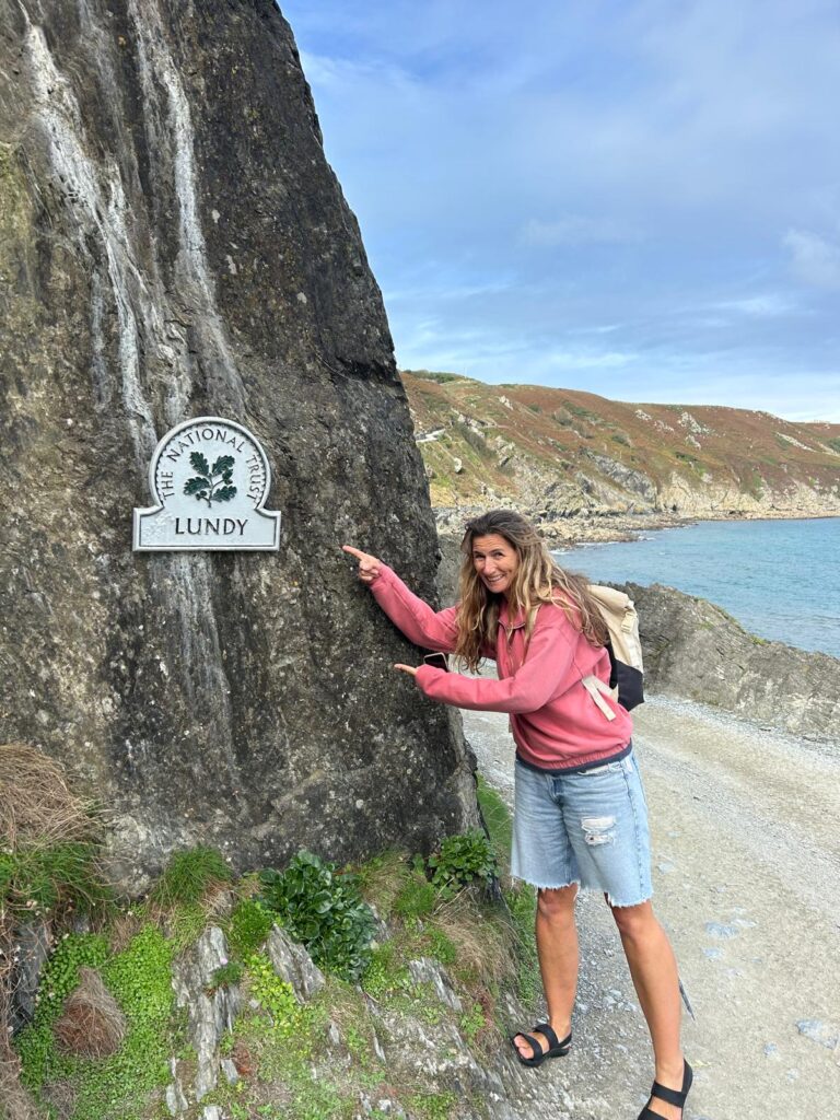 A photo of the author Ruth pointing excitedly to the Lundy Island sign on the side of a rocky path with sea behind.