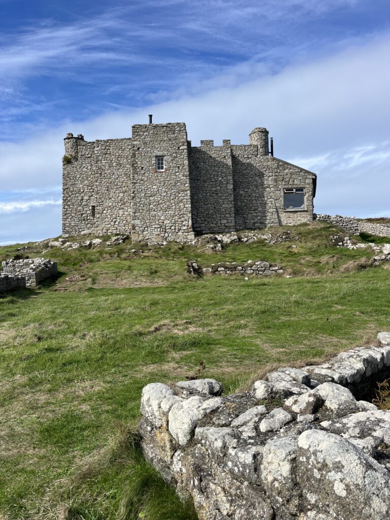 in front of a bright blue sky, a old stone house stands, surrounded by grass and small ancient stone walls.