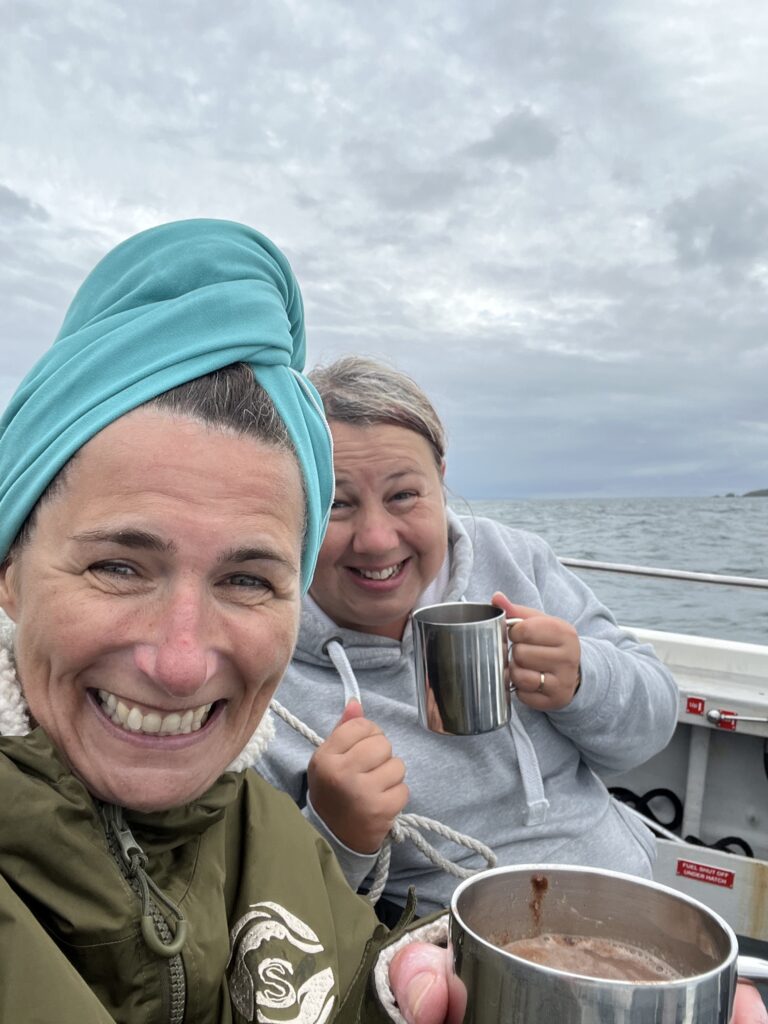 Two women sitting on the back of a boat, they are wrapped up warm after a swim and holding hot chocolates, they are both smiling widely.